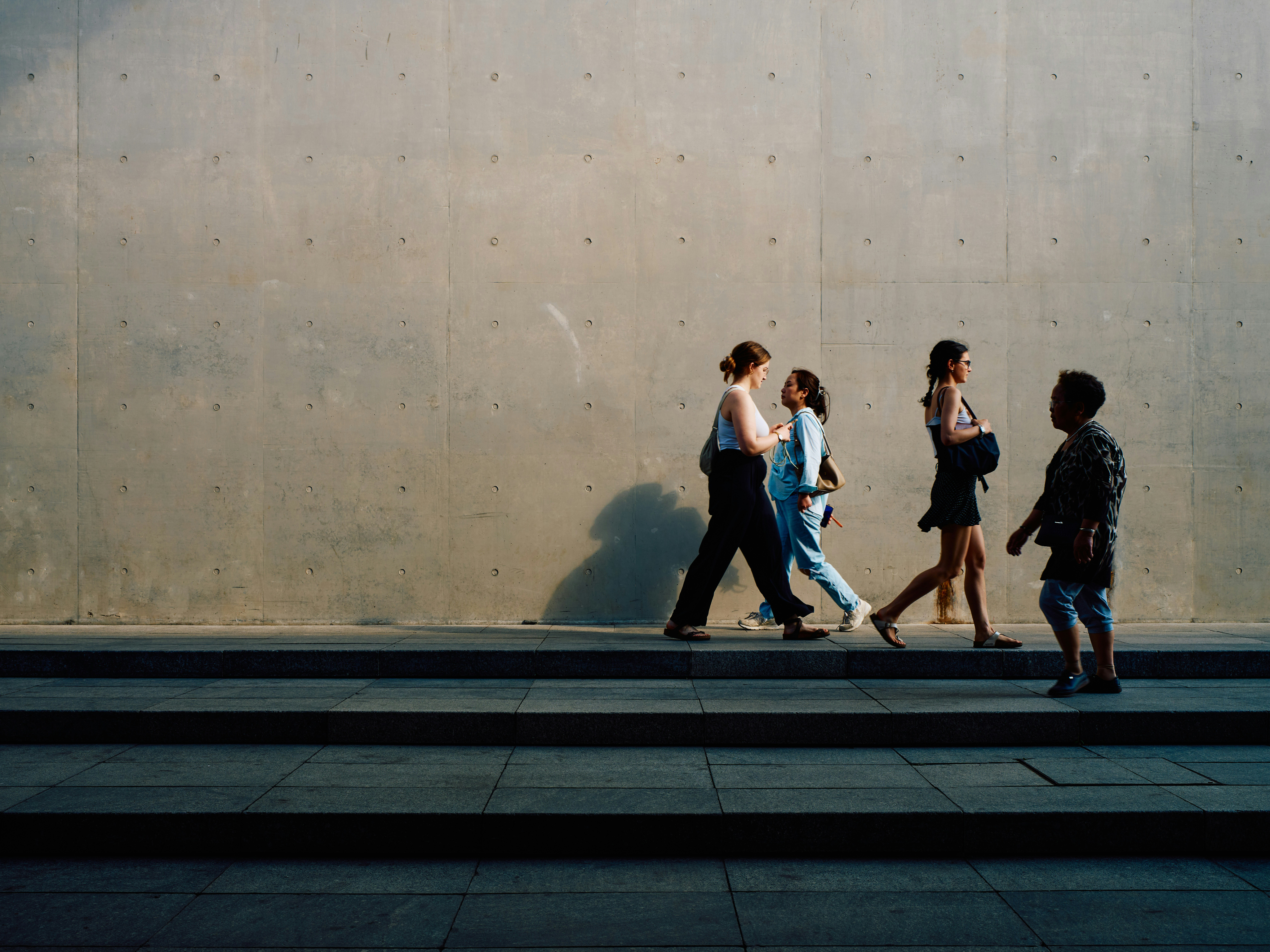 A blank concrete wall is the background to people walking past each other, getting on with their day.